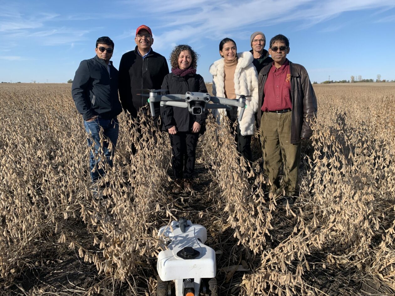 Photo of drone hovering over a farm field in Iowa with six researchers standing in the background.
