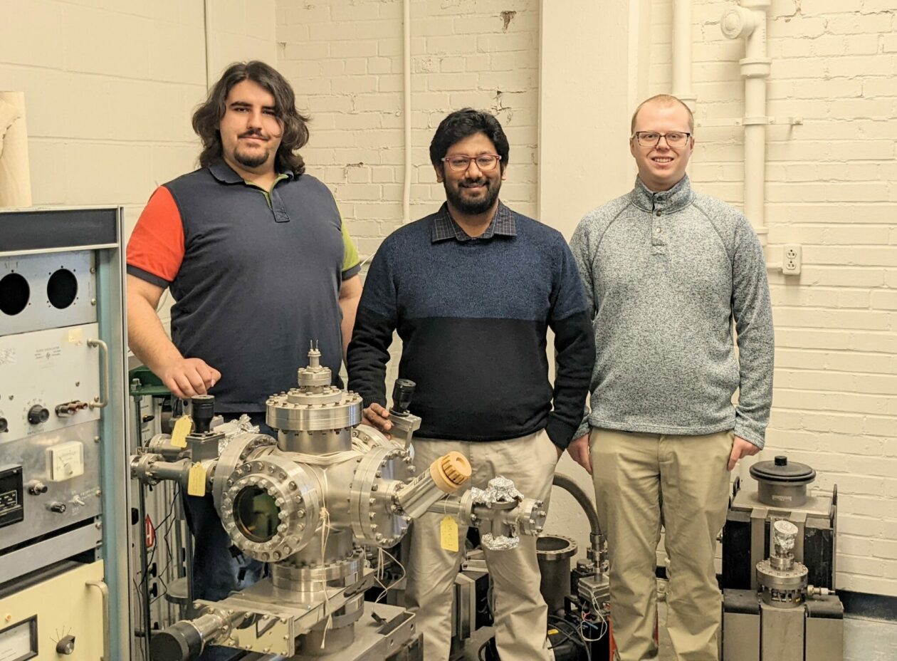 Three men stand in a nuclear engineering lab. One has long hair and a beard and wears a dark shirt with red sleeves. The man in the middle has dark hair and a beard and wears a dark shirt. The man on the right has short red hair and wear a gray shirt and tan pants.