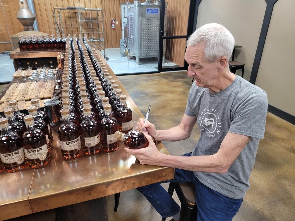 Man with short white hair, wearing a gray t-shirt, sitting at a table and signing labels on bourbon bottles.