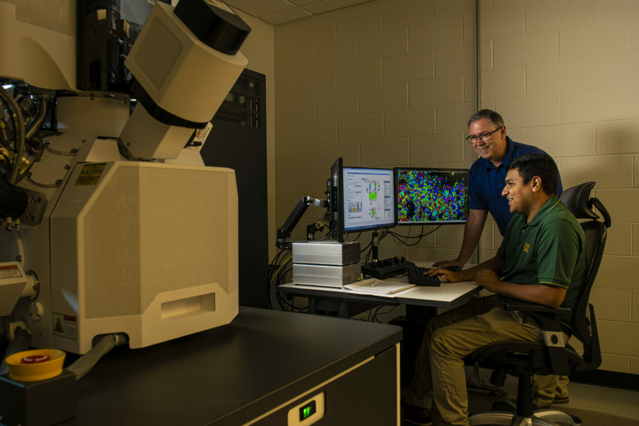 Two men working at a computer. The man standing is wearing glasses and a blue polo shirt. The seated man is searing a green polo and khaki pants. The computer displays a colorful X-ray image.