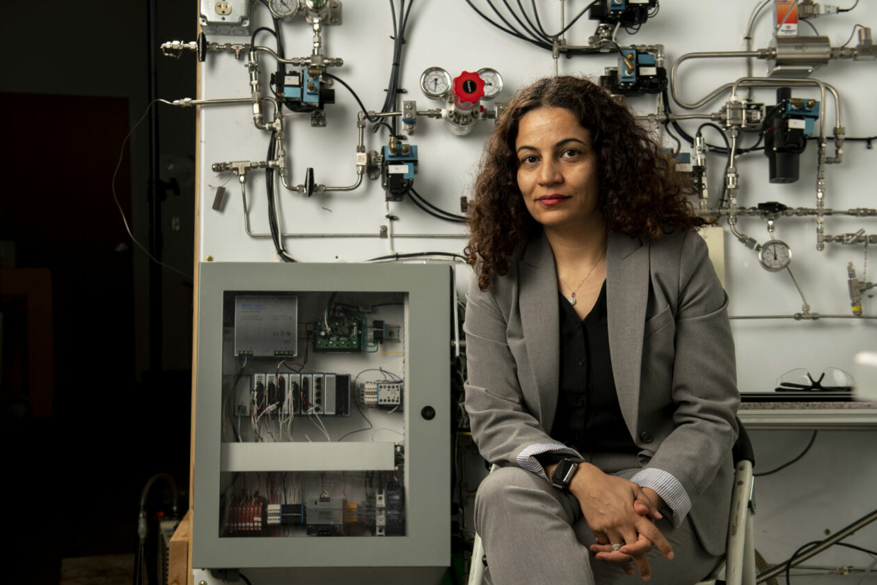 Woman with long, dark, curly hair wearing a tan suit and black top sitting in front of lab equipment