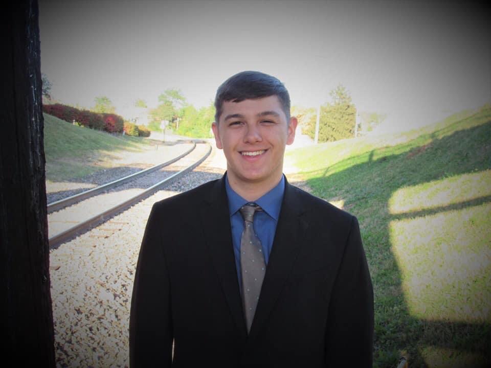 young man in dark suit, blue shirt and gray tie stands outside