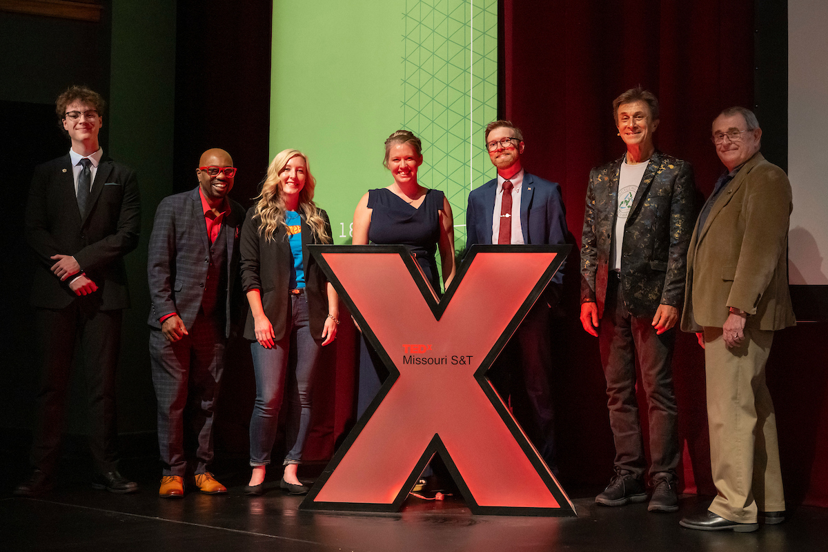 Seven adults stand by large red X that symbolizes TEDx