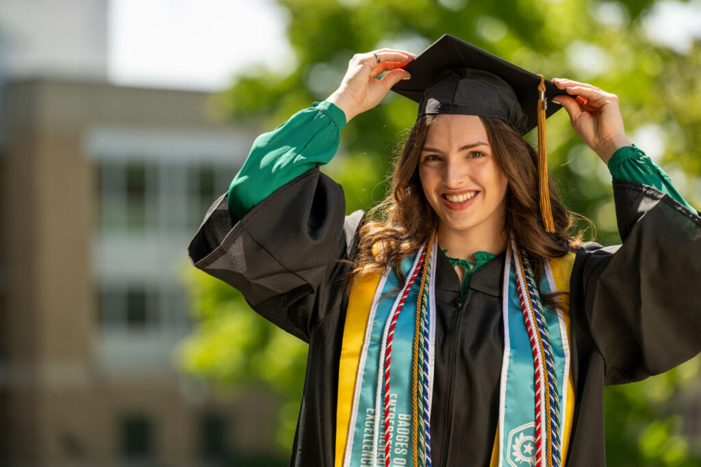 Woman in academic regalia adjusts graduation cap. 