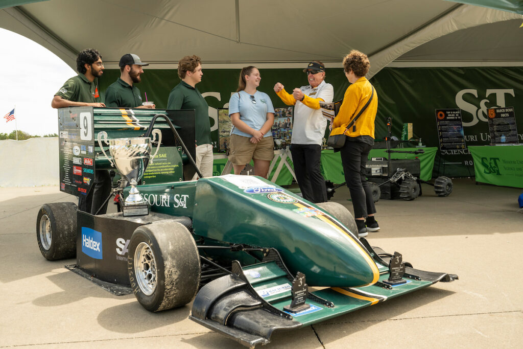 Three students, one staff member and two adults near a race car with a trophy on display