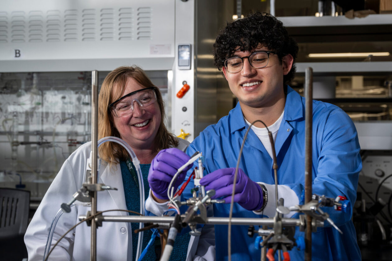 Dr. Minteer watches as student uses lab equipment. Both wear goggles and lab coats.