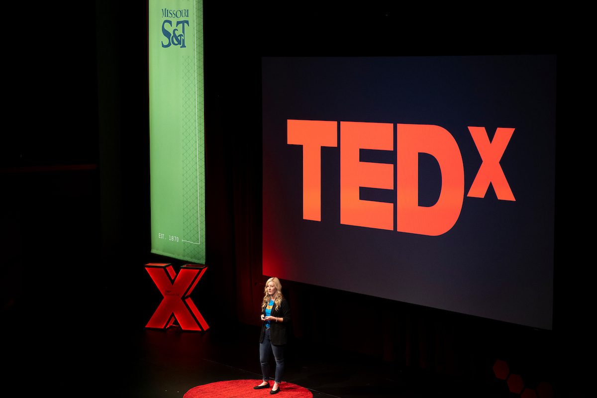 A blonde woman stands on a red circle on a dark stage. Behind her, a screen shows the TEDx logo in red, there is a green banner with the Missouri S&T logo, and a decorative red X sits on the stage.